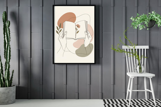 Hands holding an open book with a pressed flower, line art print framed in a room with plants