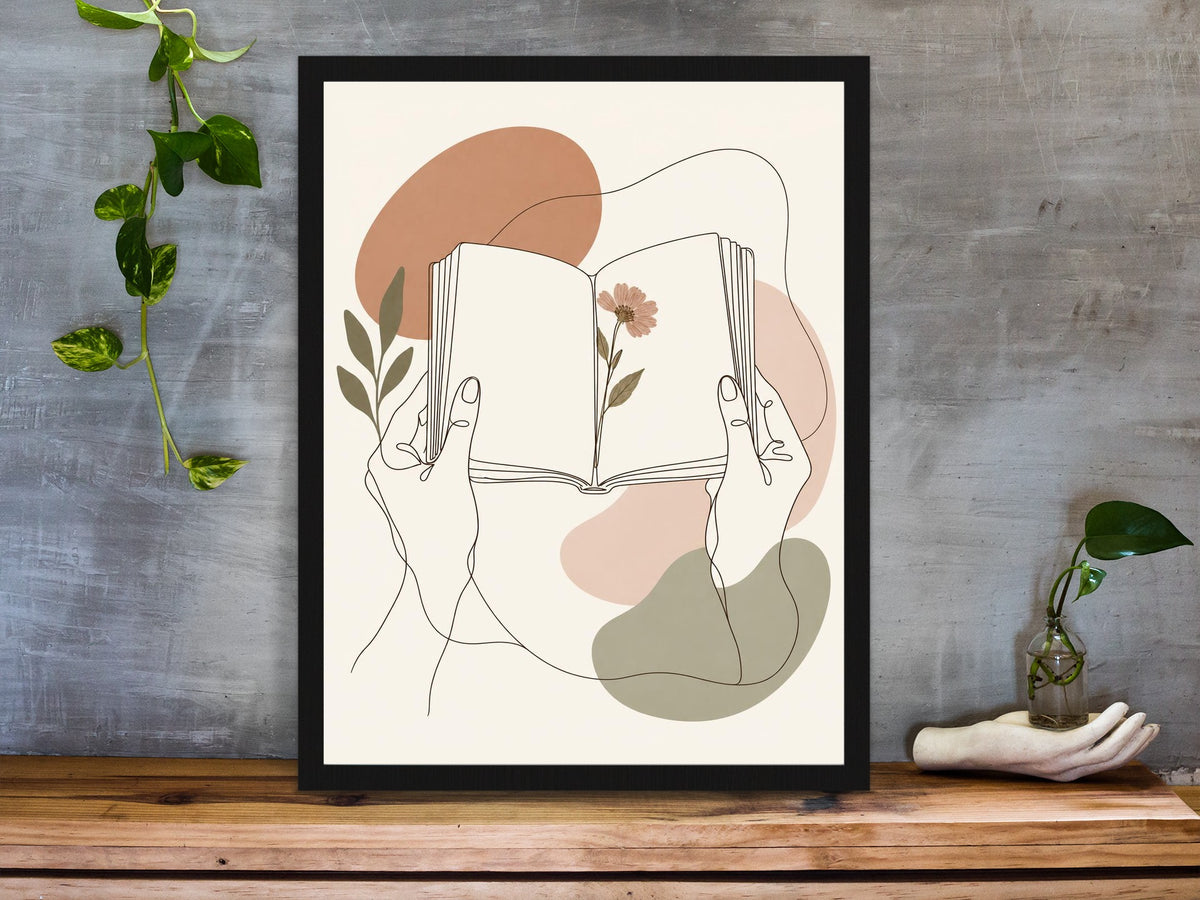 Hands holding an open book with a pressed flower, line art print framed on a desk