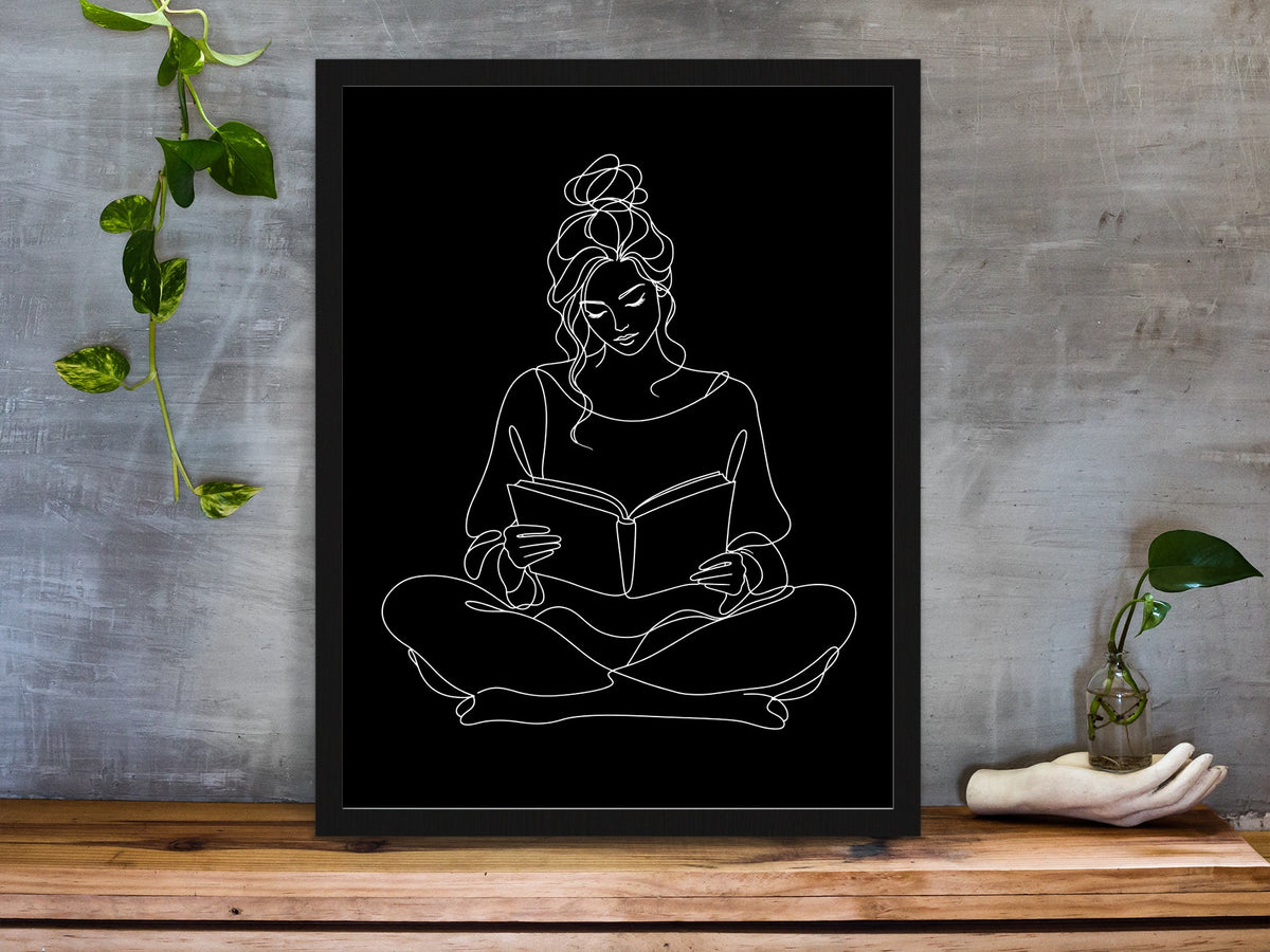 Woman sitting cross-legged reading a book, continuous line art print, white lines on black, framed on a desk