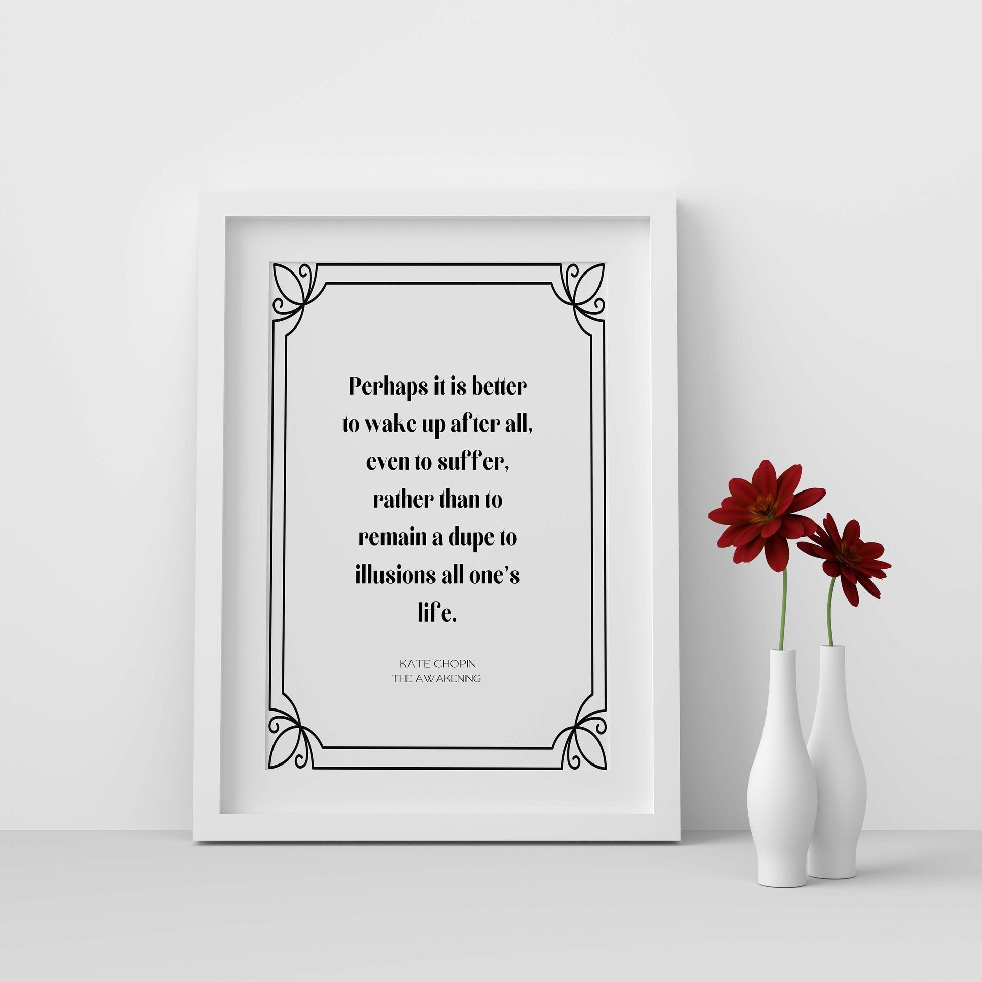 A black and white framed print with a quote by Kate Chopin from 'The Awakening' that reads: 'Perhaps it is better to wake up after all, even to suffer, rather than to remain a dupe to illusions all one's life.' The print is displayed on a white wall with two red flowers in white vases in the foreground.
