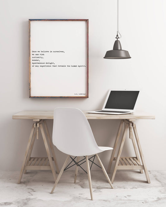 May include: A framed quote on a white wall with a black lamp hanging above it. The quote reads 'Once we believe in ourselves, we can risk curiosity, wonder, spontaneous delight, or any experience that reveals the human spirit.' The quote is attributed to 'E.E. Cummings.' A white chair and a laptop are on a wooden desk in front of the wall.