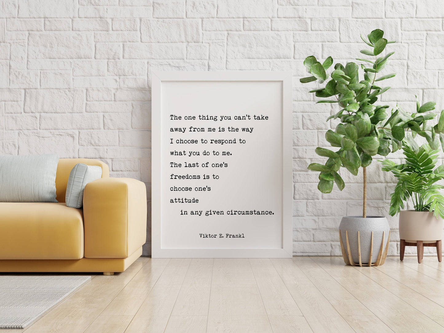 May include: A white framed print with a black and white quote by Viktor E. Frankl that reads 'The one thing you can't take away from me is the way I choose to respond to what you do to me. The last of one's freedoms is to choose one's attitude in any given circumstance. Viktor E. Frankl' The print is hanging on a white brick wall. A yellow couch is in the foreground. A green plant in a gray pot with a brown stand is on the right side of the image.