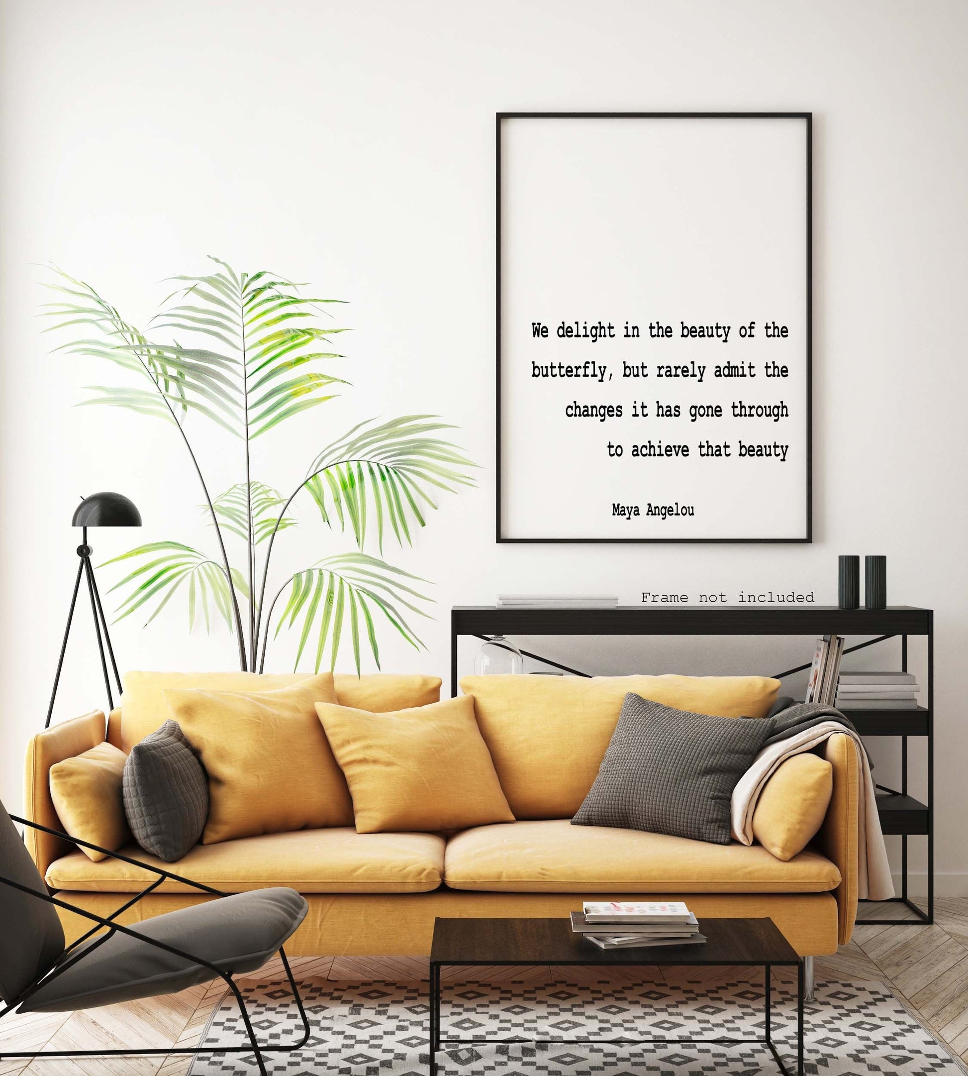 May include: A black framed print with a white background and black text that reads 'We delight in the beauty of the butterfly, but rarely admit the changes it has gone through to achieve that beauty Maya Angelou Frame not included' The print is hanging on a white wall above a yellow couch with black throw pillows. There is a green plant in a pot to the left of the print.