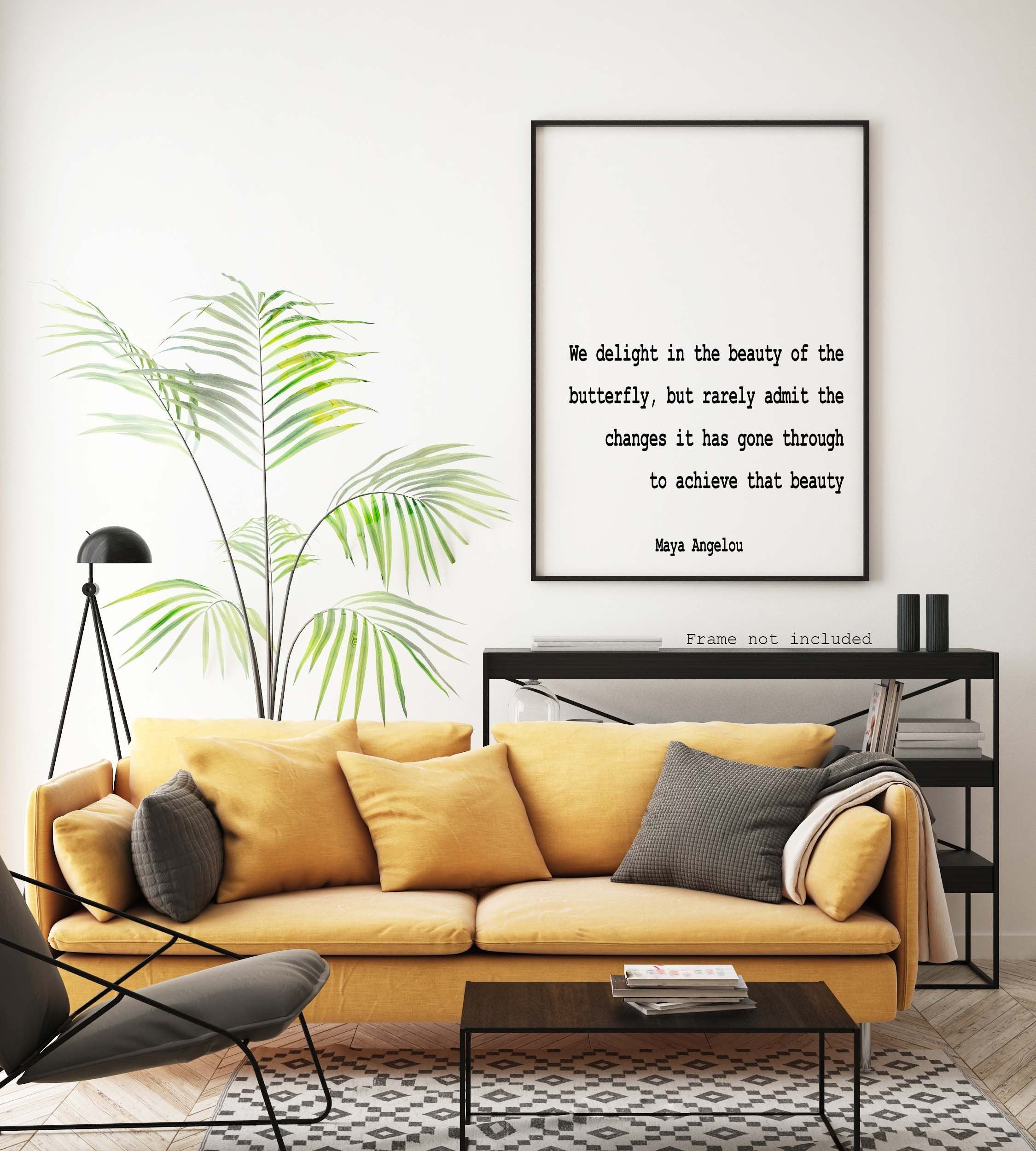 May include: A black framed print with a white background and black text that reads 'We delight in the beauty of the butterfly, but rarely admit the changes it has gone through to achieve that beauty Maya Angelou Frame not included' The print is hanging on a white wall above a yellow couch with black throw pillows. There is a green plant in a pot to the left of the print.