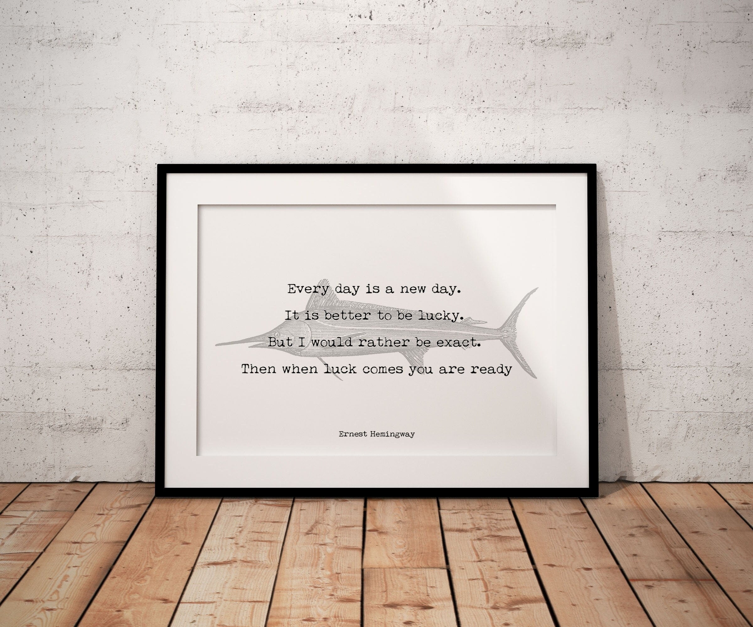 May include: A black framed print with a white background. The print features a quote by Ernest Hemingway: 'Every day is a new day. It is better to be lucky. But I would rather be exact. Then when luck comes you are ready.' The quote is printed in a typewriter font and is centered on the page. A stylized image of a marlin is behind the quote.