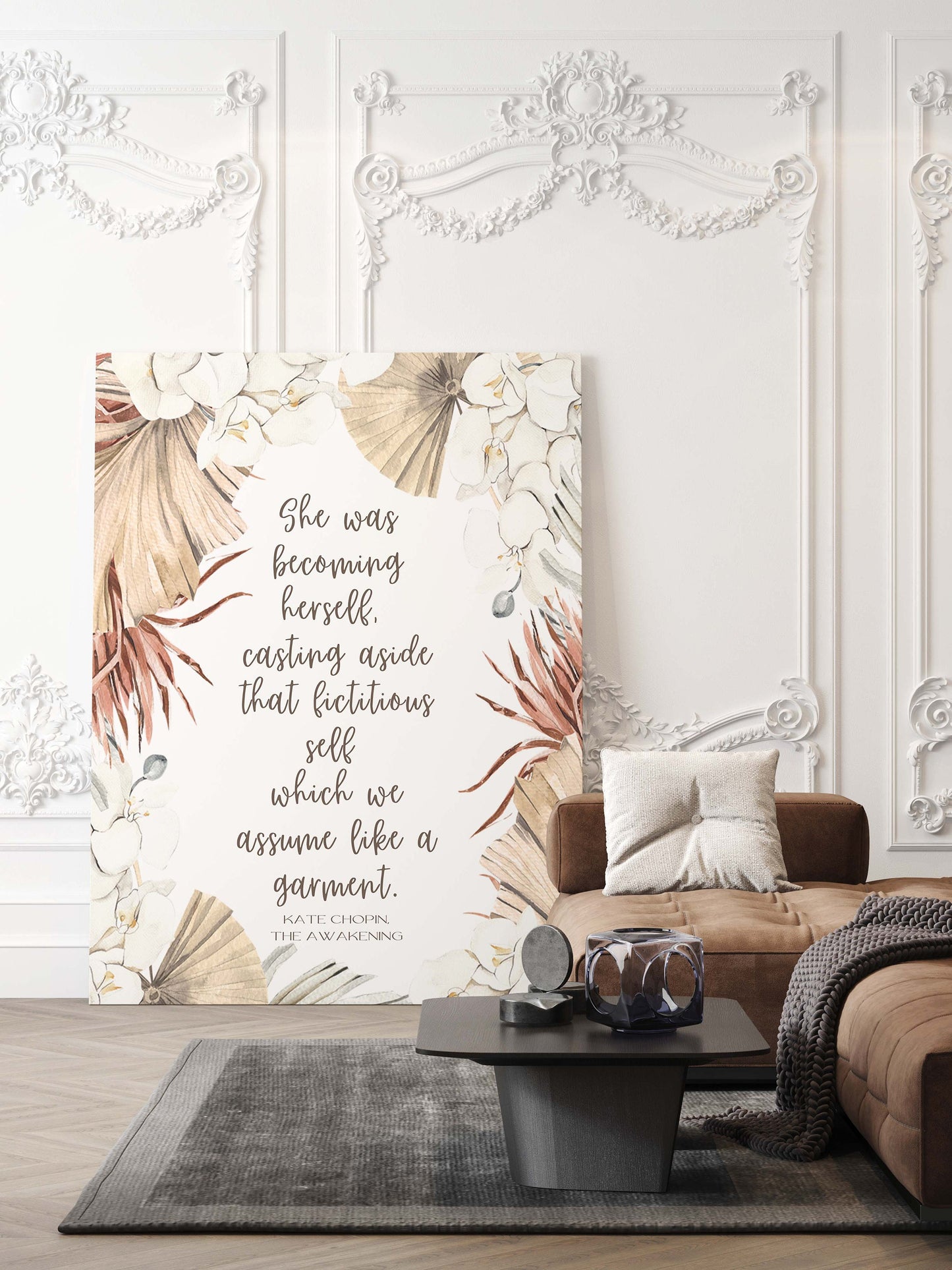 May include: A canvas print with a floral design and a quote from Kate Chopin's The Awakening: 'She was becoming herself. Casting aside that fictitious self which we assume like a garment.' The quote is printed in white text on a beige background. The floral design features white flowers, brown palm leaves, and beige leaves. The canvas print is hanging on a white wall with ornate molding.