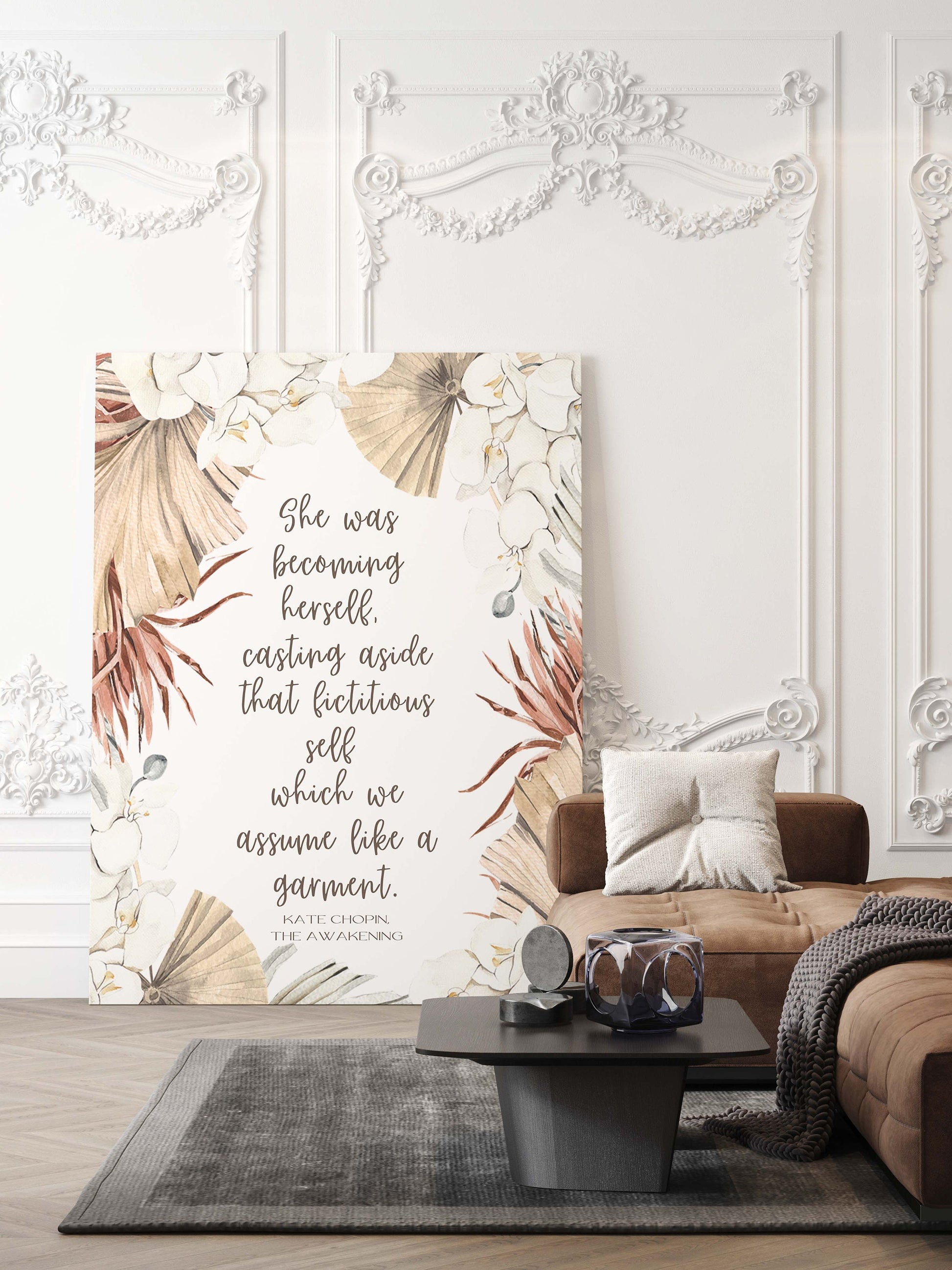 May include: A canvas print with a floral design and a quote from Kate Chopin's The Awakening: 'She was becoming herself. Casting aside that fictitious self which we assume like a garment.' The quote is printed in white text on a beige background. The floral design features white flowers, brown palm leaves, and beige leaves. The canvas print is hanging on a white wall with ornate molding.