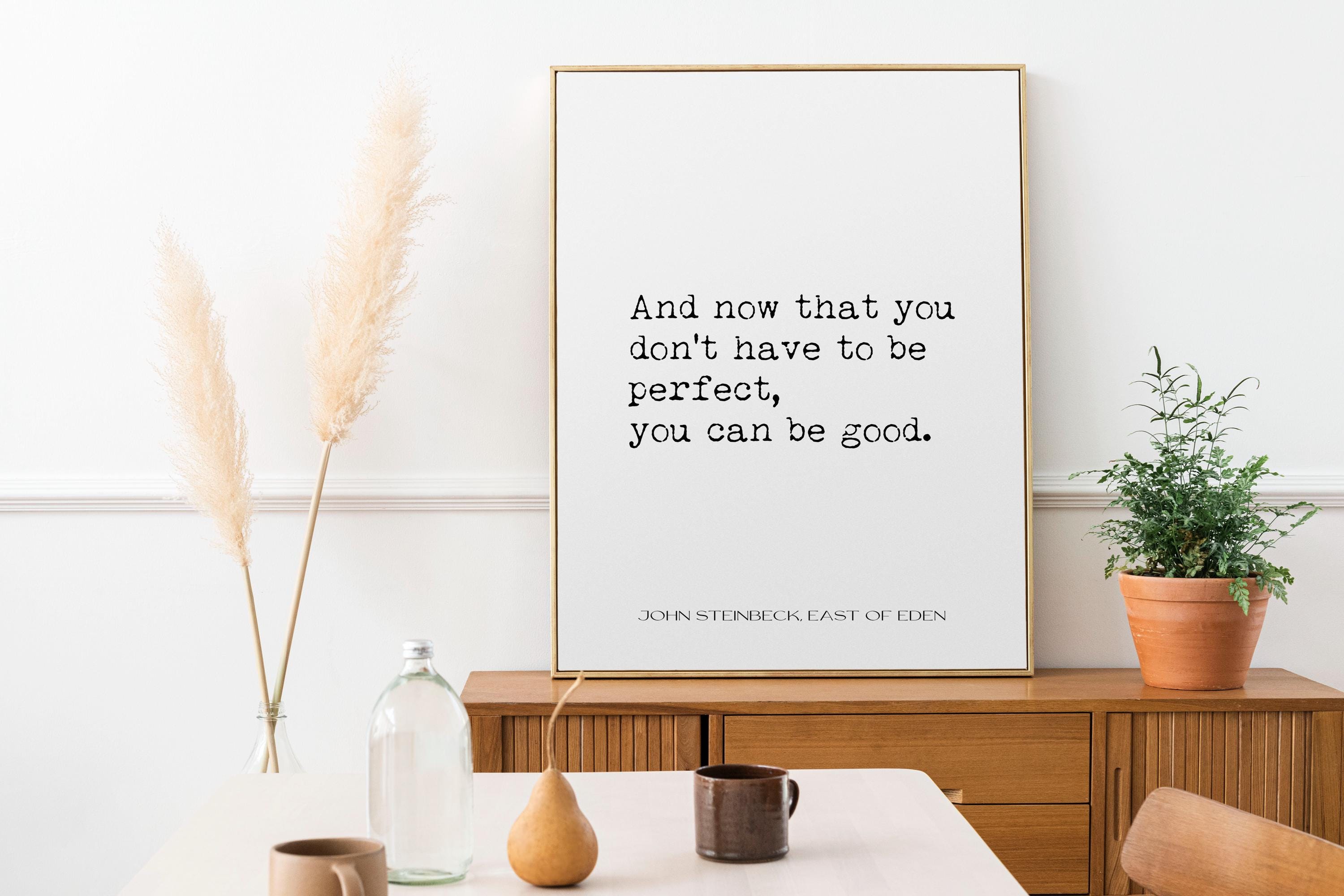 May include: A framed print with a white background and black text that reads 'And now that you don't have to be perfect, you can be good. John Steinbeck East of Eden' The print is hanging on a white wall above a wooden dresser. There are two tall dried pampas grass plants in clear glass vases on the dresser. There is a small potted plant on the right side of the dresser.