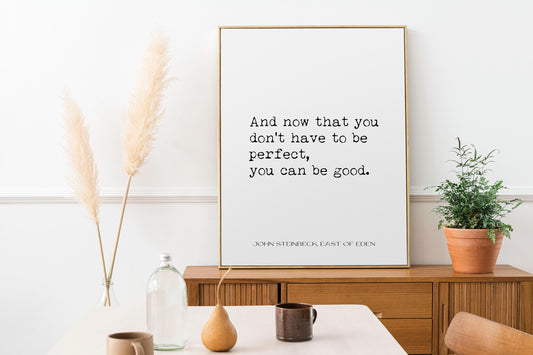 May include: A framed print with a white background and black text that reads 'And now that you don't have to be perfect, you can be good. John Steinbeck East of Eden' The print is hanging on a white wall above a wooden dresser. There are two tall dried pampas grass plants in clear glass vases on the dresser. There is a small potted plant on the right side of the dresser.