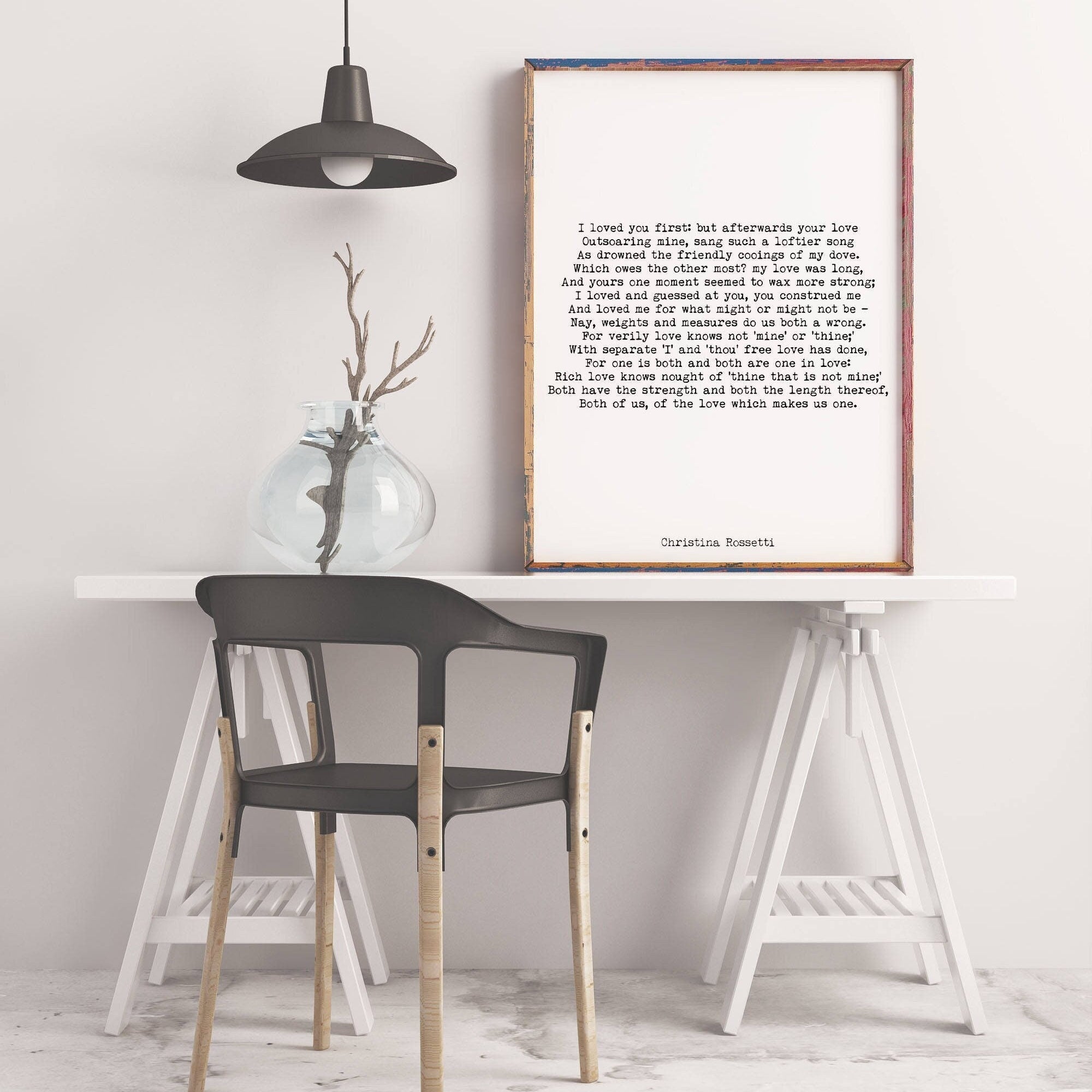 May include: A black and white framed print with a poem titled 'I loved you first' by Christina Rossetti. The poem is printed in a typewriter font. The print is hanging on a white wall above a white table with a black chair and a white easel.