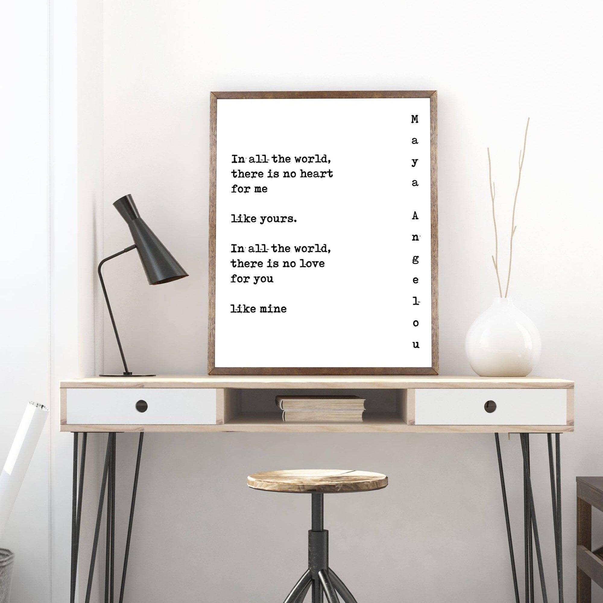 May include: A framed black and white typography print with the text 'In all the world, there is no heart for me like yours. In all the world, there is no love for you like mine.' The print is hanging on a white wall above a wooden desk with a white drawer and a black lamp. A white vase with a branch is on the right side of the desk. A black stool is in front of the desk.