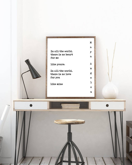 May include: A framed black and white typography print with the text 'In all the world, there is no heart for me like yours. In all the world, there is no love for you like mine.' The print is hanging on a white wall above a wooden desk with a white drawer and a black lamp. A white vase with a branch is on the right side of the desk. A black stool is in front of the desk.