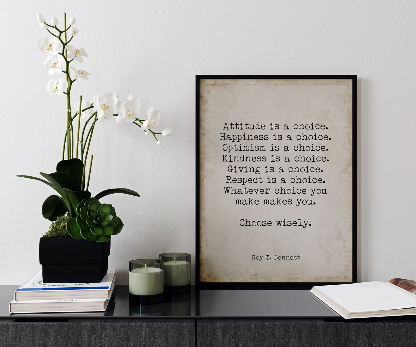 May include: A black framed print with a beige background and white text that reads: 'Attitude is a choice. Happiness is a choice. Optimism is a choice. Kindness is a choice. Giving is a choice. Respect is a choice. Whatever choice you make makes you. Choose wisely. Roy T. Bennett' The print is sitting on a black surface with a white orchid plant in a black planter and two glass candle holders with green candles.