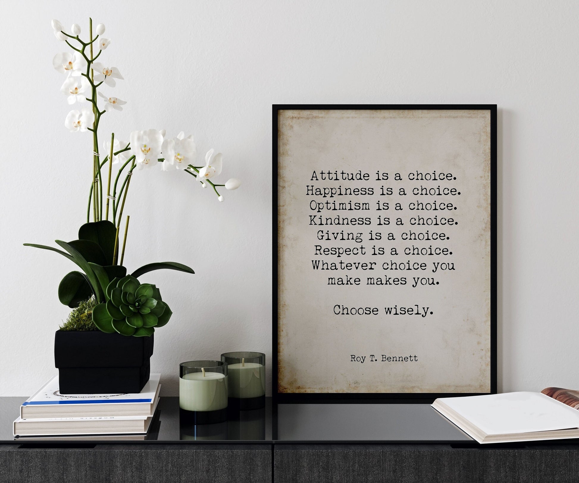May include: A black framed print with a beige background and white text that reads: 'Attitude is a choice. Happiness is a choice. Optimism is a choice. Kindness is a choice. Giving is a choice. Respect is a choice. Whatever choice you make makes you. Choose wisely. Roy T. Bennett' The print is sitting on a black surface with a white orchid plant in a black planter and two glass candle holders with green candles.
