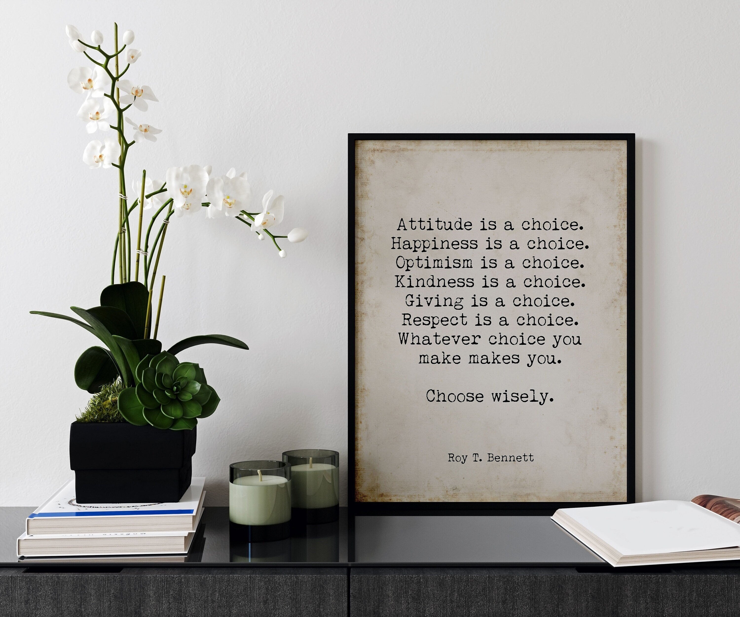 May include: A black framed print with a beige background and white text that reads: 'Attitude is a choice. Happiness is a choice. Optimism is a choice. Kindness is a choice. Giving is a choice. Respect is a choice. Whatever choice you make makes you. Choose wisely. Roy T. Bennett' The print is sitting on a black surface with a white orchid plant in a black planter and two glass candle holders with green candles.