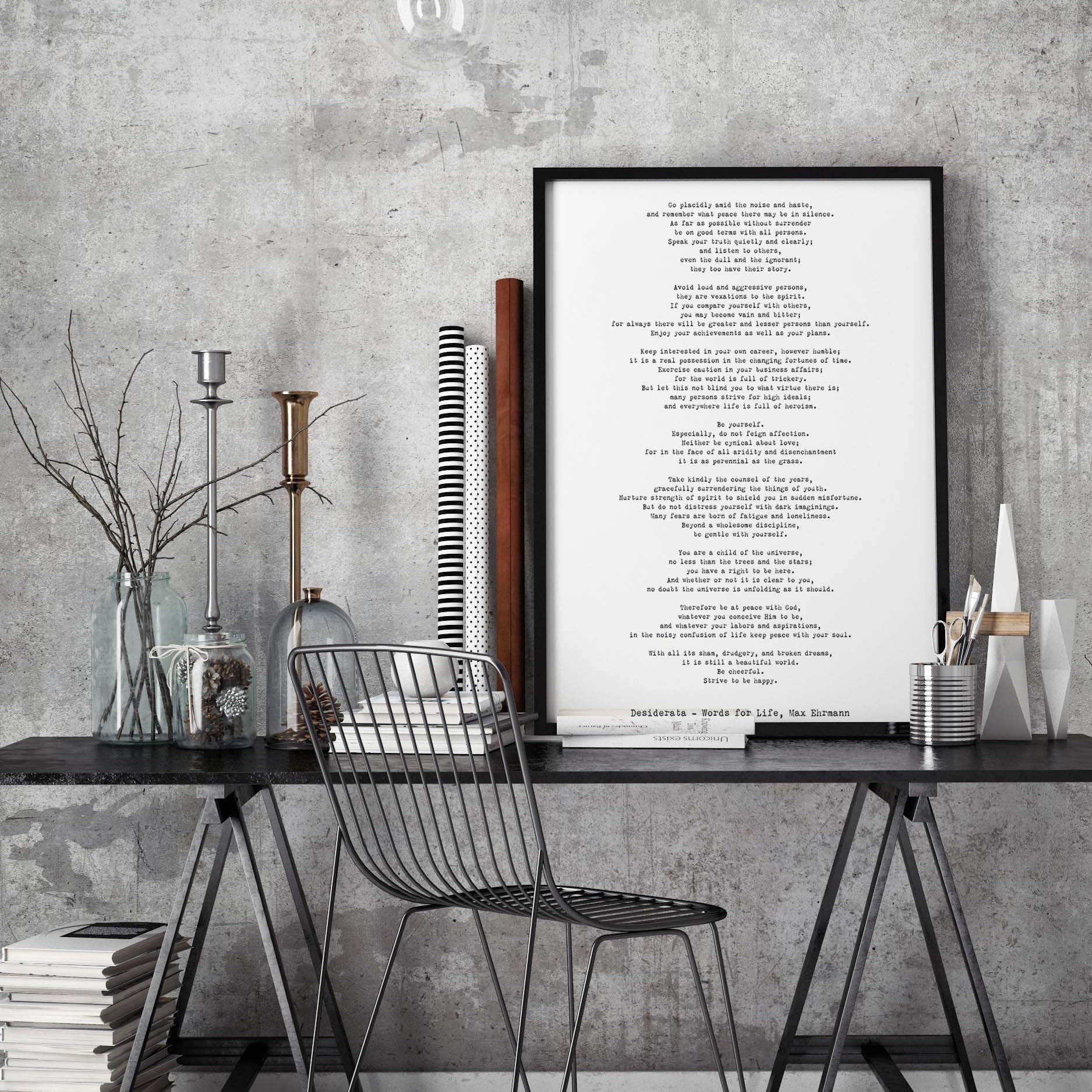 May include: A black metal chair with a wire frame design sits in front of a black wooden desk with a black metal frame. The desk has a stack of books and a silver metal container with white paintbrushes. A framed black and white print with the text 'Decalogue - Words For Life, Max Ehrmann' hangs on a gray concrete wall behind the desk. A black metal light fixture hangs above the desk.
