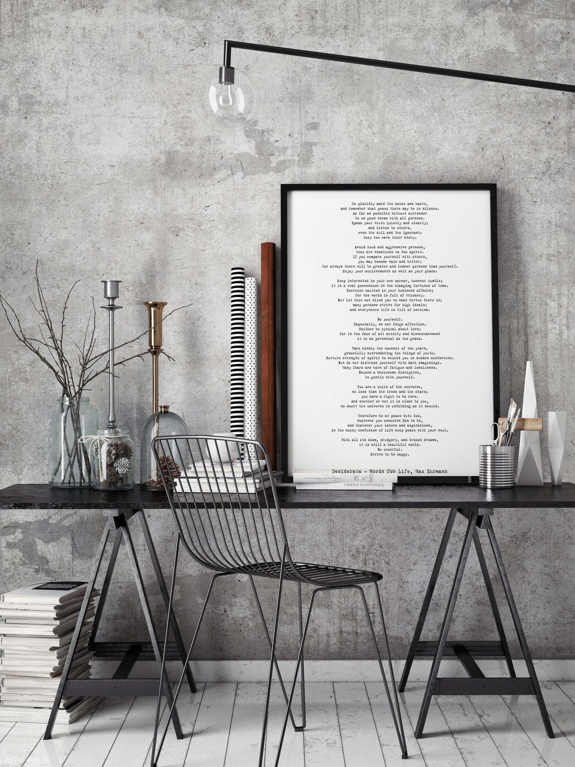 May include: A black metal chair with a wire frame design sits in front of a black wooden desk with a black metal frame. The desk has a stack of books and a silver metal container with white paintbrushes. A framed black and white print with the text 'Decalogue - Words For Life, Max Ehrmann' hangs on a gray concrete wall behind the desk. A black metal light fixture hangs above the desk.