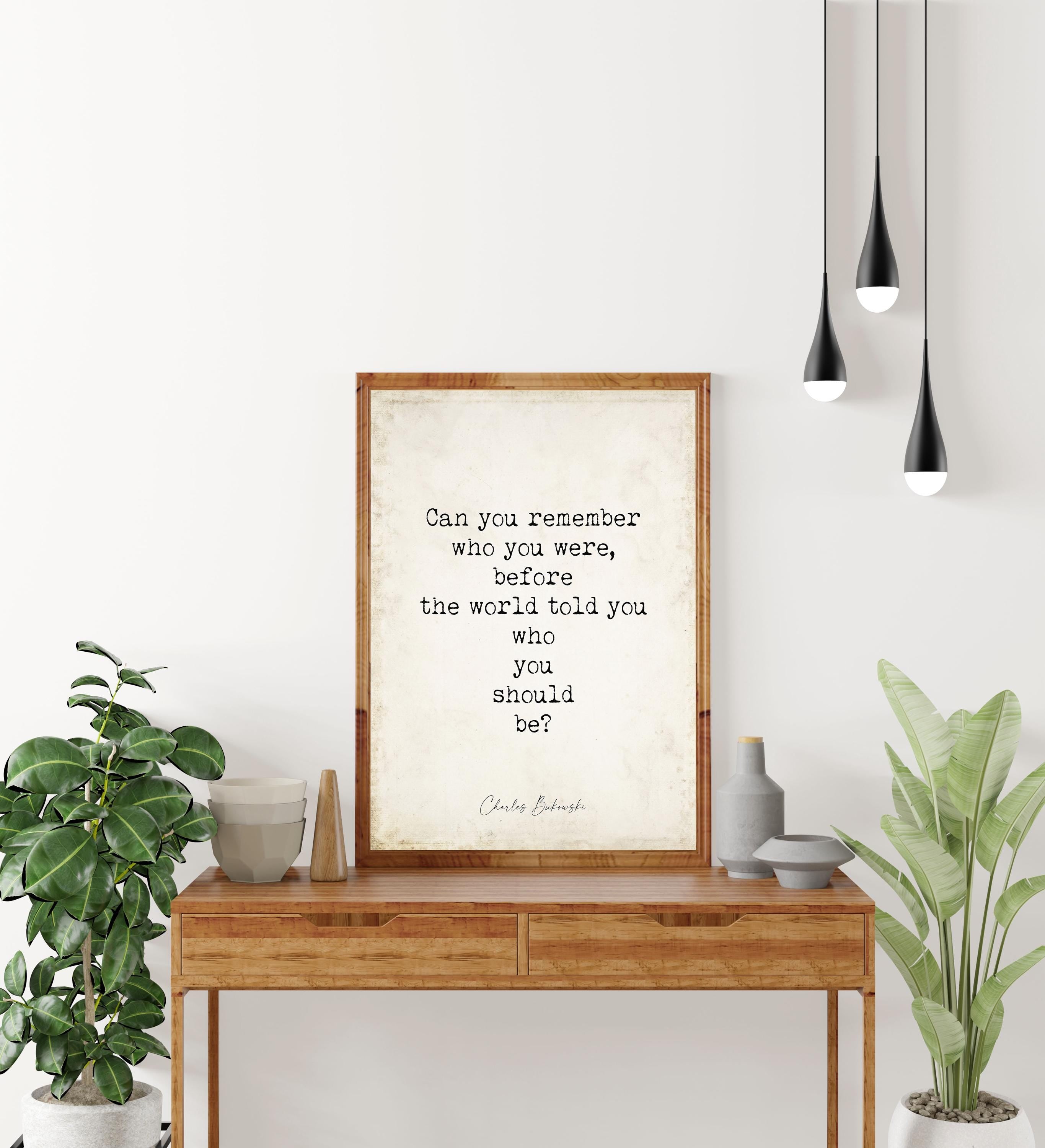 May include: A framed print with a quote on a wooden table. The quote reads, 'Can you remember who you were, before the world told you who you should be?' by Charles Bukowski. The table has two drawers and is surrounded by plants and decorative items. Three black pendant lights hang above.