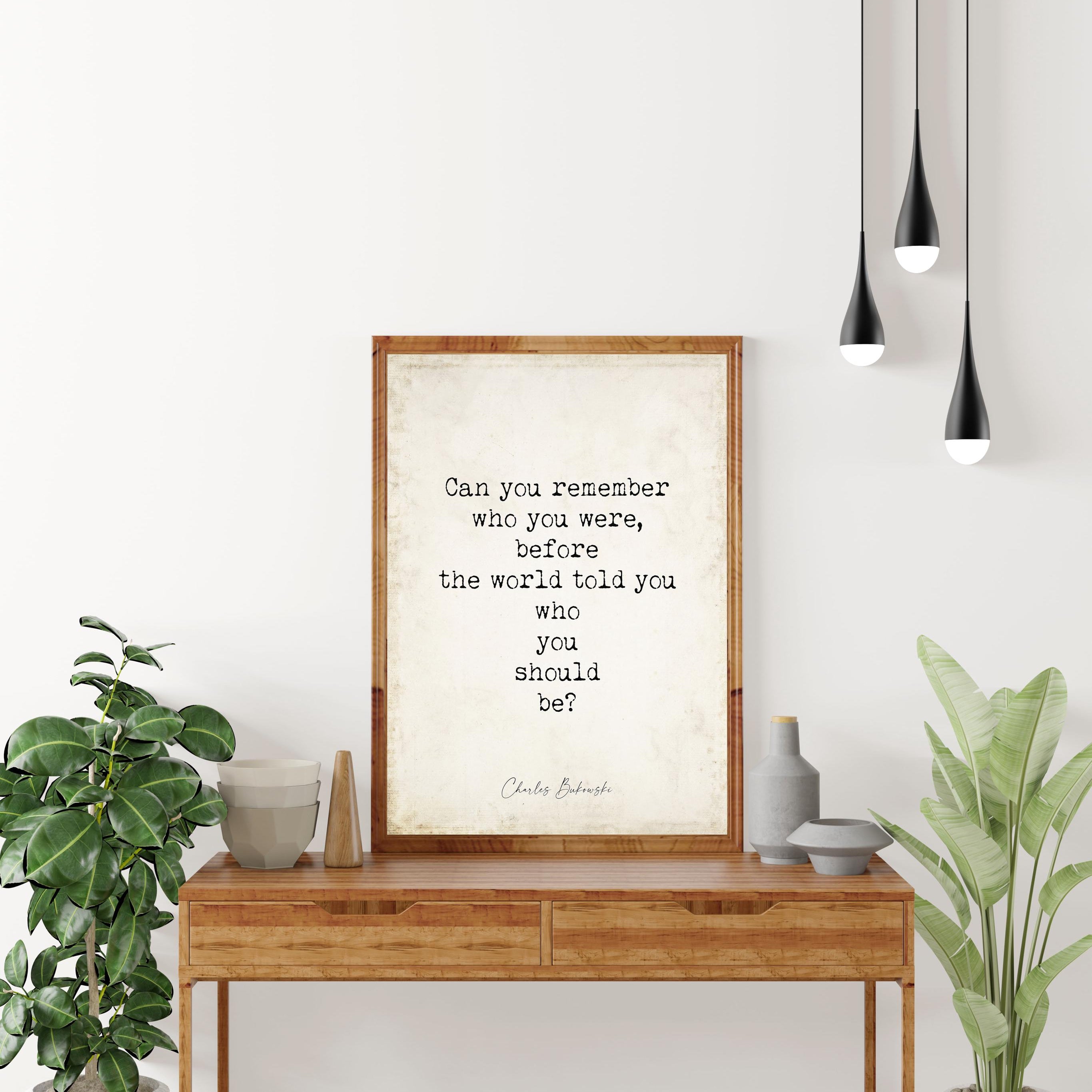 May include: A framed print with a quote on a wooden table. The quote reads, 'Can you remember who you were, before the world told you who you should be?' by Charles Bukowski. The table has two drawers and is surrounded by plants and decorative items. Three black pendant lights hang above.