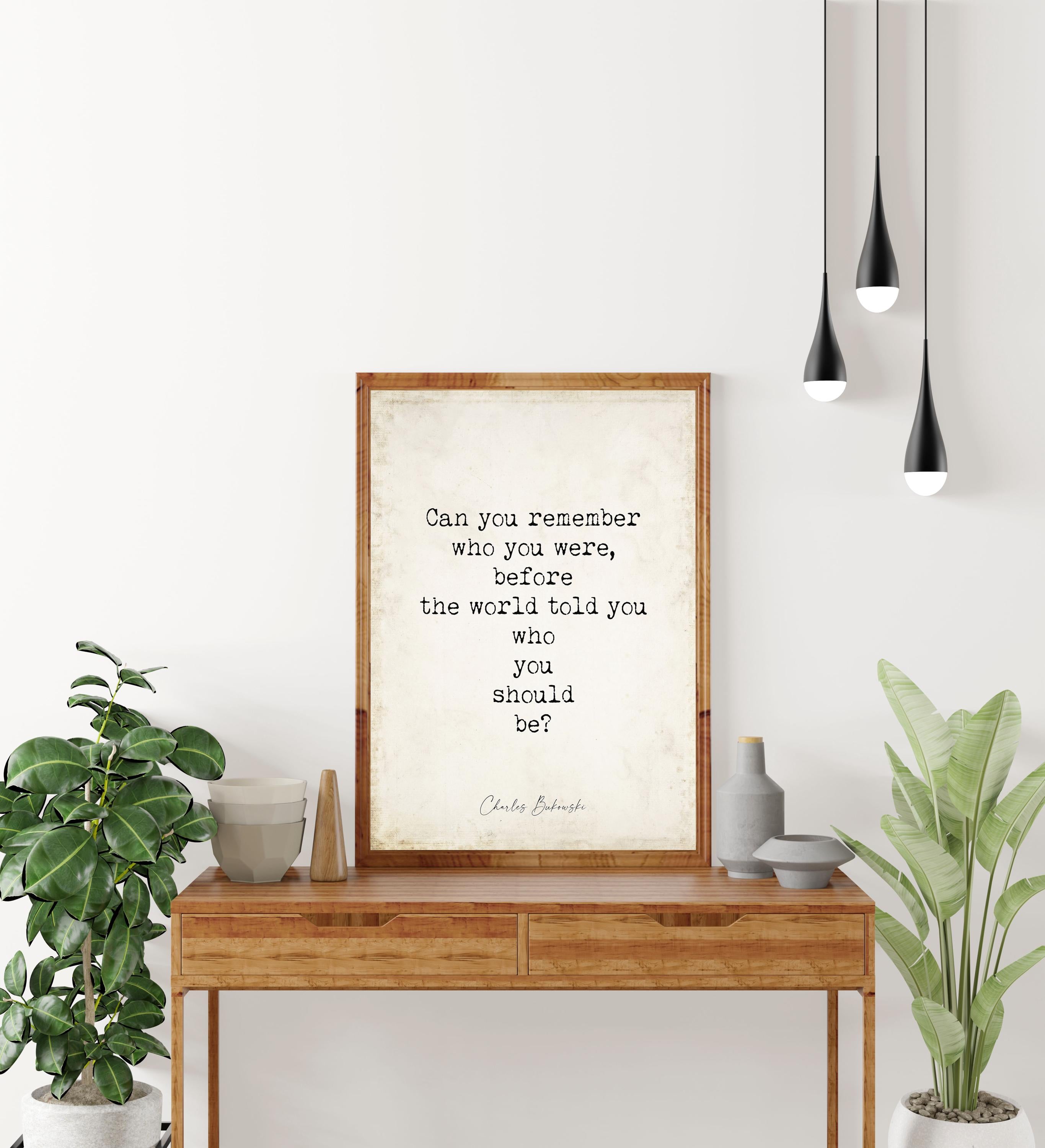 May include: A framed print with a quote on a wooden table. The quote reads, 'Can you remember who you were, before the world told you who you should be?' by Charles Bukowski. The table has two drawers and is surrounded by plants and decorative items. Three black pendant lights hang above.