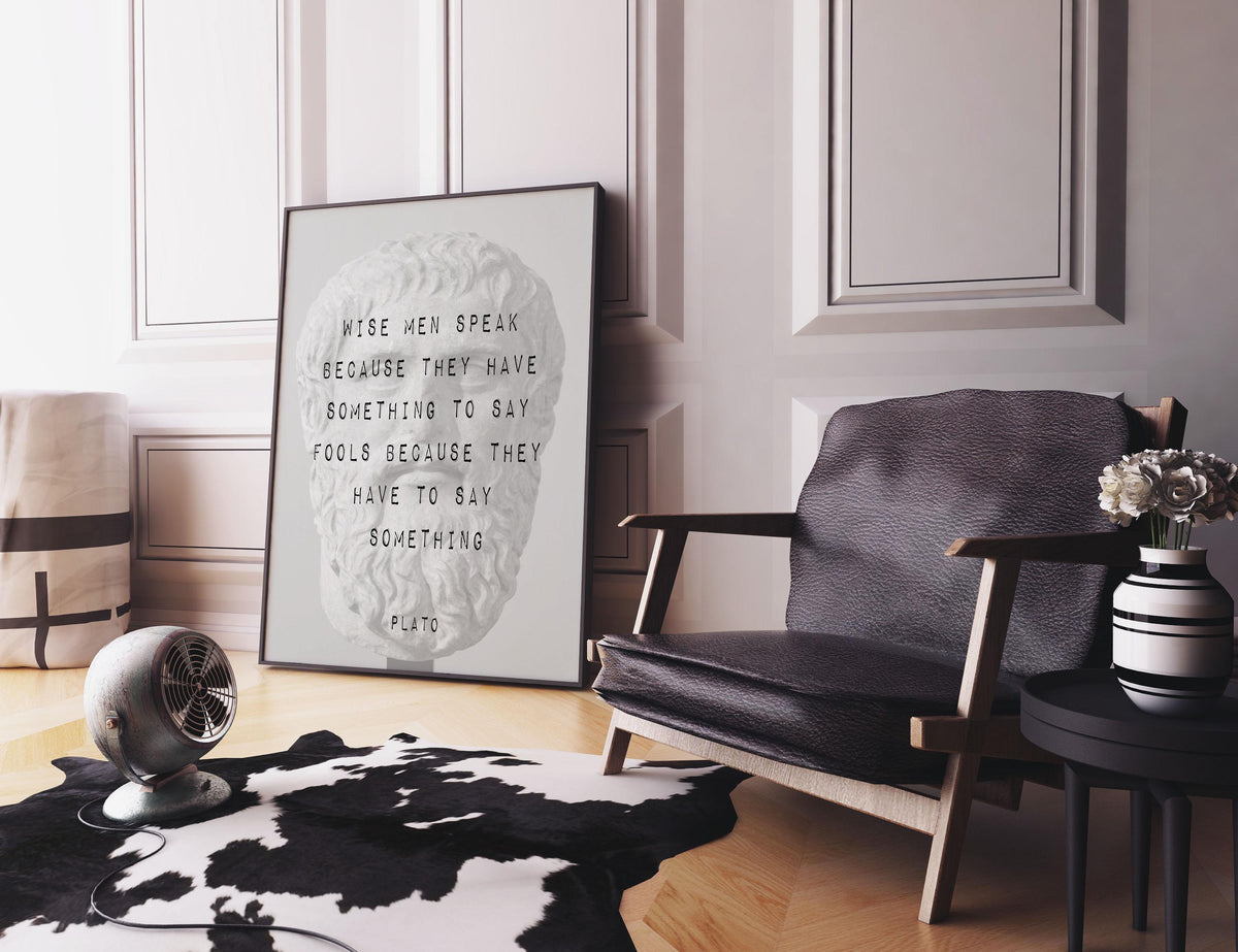 A framed print of a Plato bust with a quote is on a black and white cowhide rug next to a black leather armchair, a small round table with a vase of flowers, and a metal fan.