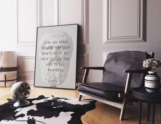 A framed print of a Plato bust with a quote is on a black and white cowhide rug next to a black leather armchair, a small round table with a vase of flowers, and a metal fan.