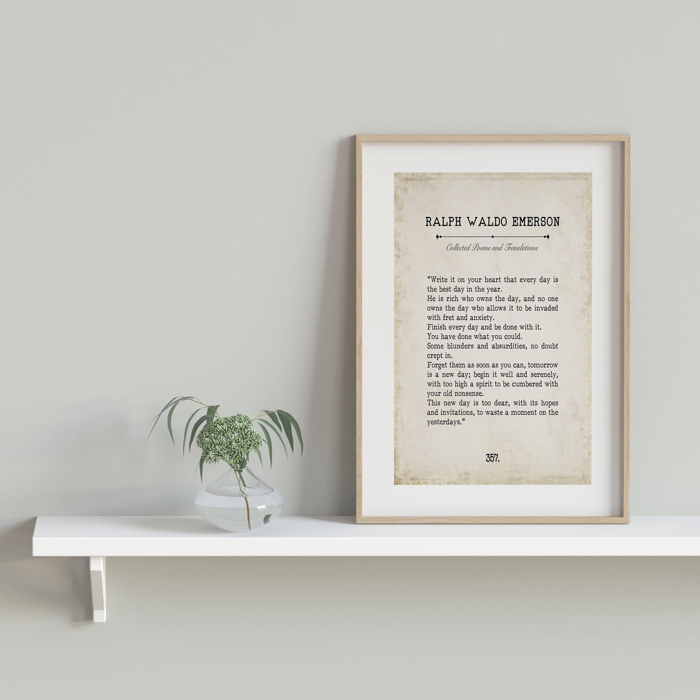 A framed quote print, titled "Collected Poems and Translations" by Ralph Waldo Emerson, sits on a white shelf with a small glass vase containing a green plant.