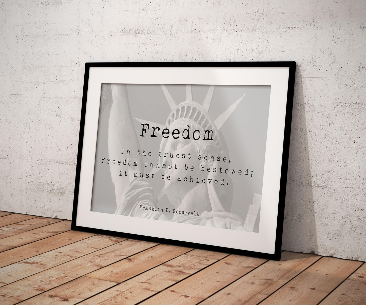 A framed print with the word "Freedom" and a quote by Franklin D. Roosevelt, overlaid on a faded image of the Statue of Liberty, leaning against a textured white wall on a wooden floor.