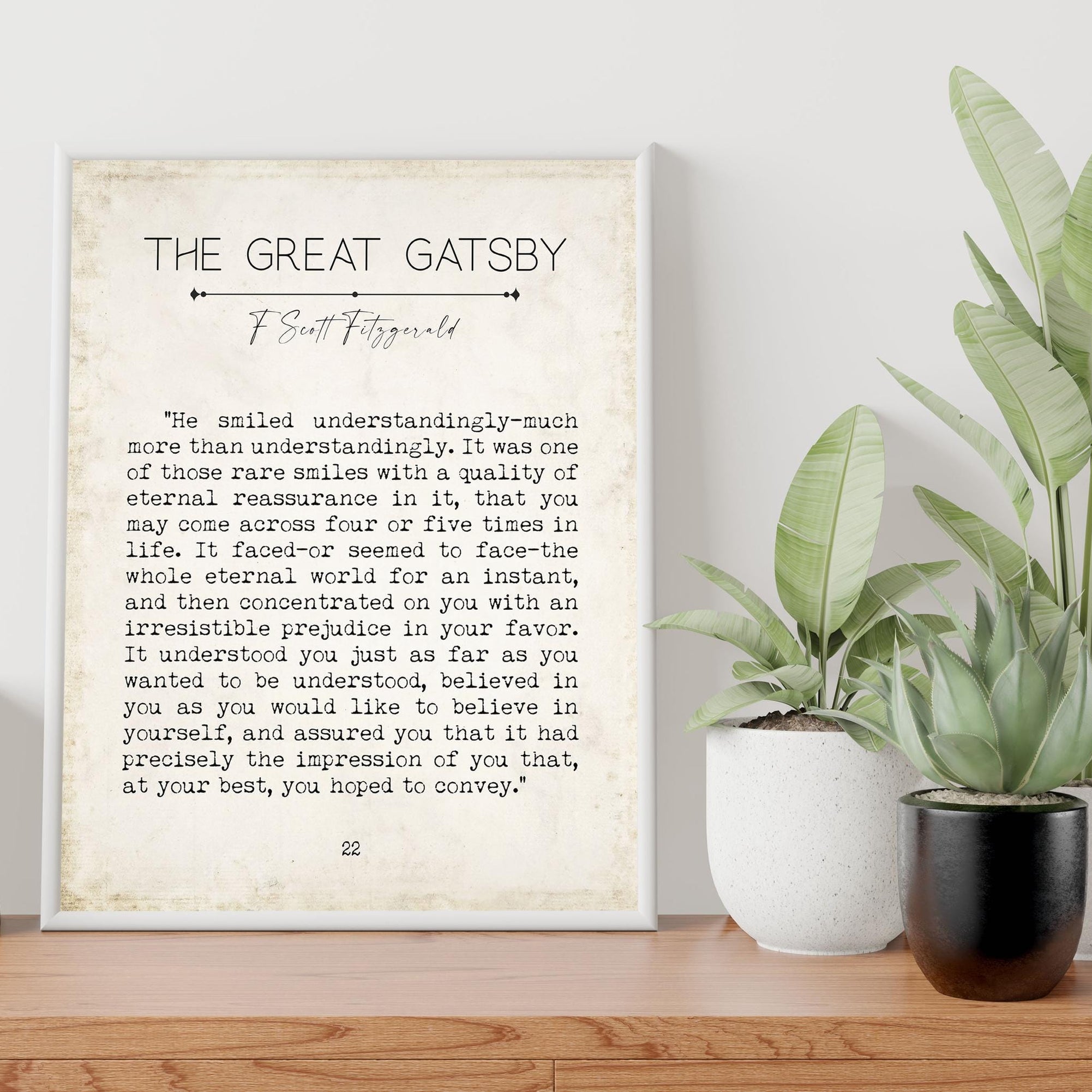 A framed print of a "The Great Gatsby" quote on a light wooden surface, with two potted plants to the right, against a light gray wall.