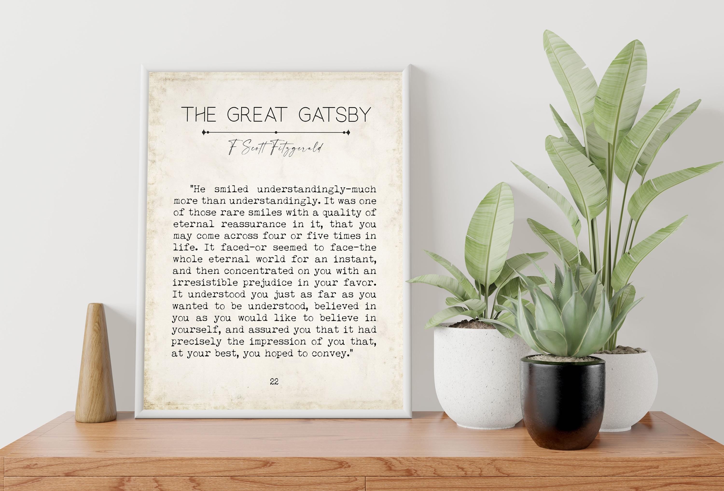 A framed print of a "The Great Gatsby" quote on a light wooden surface, with two potted plants to the right, against a light gray wall.