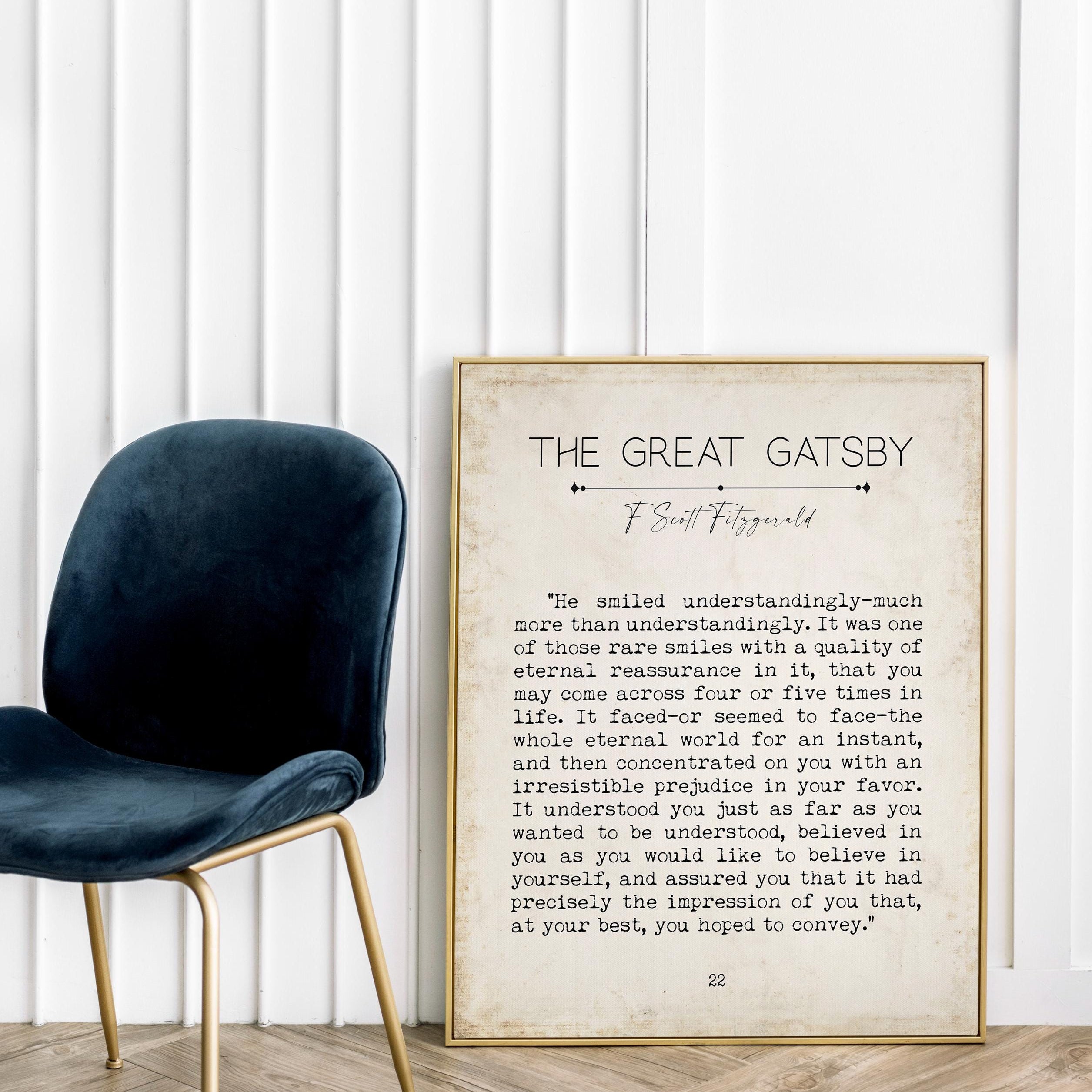 A framed print of a "The Great Gatsby" quote on the floor next to a dark blue velvet chair, against a white wall.