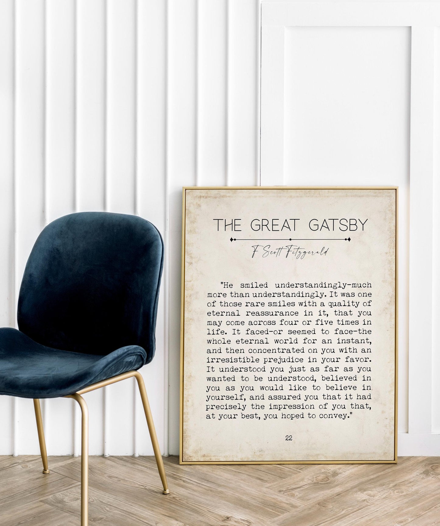 A framed print of a "The Great Gatsby" quote on the floor next to a dark blue velvet chair, against a white wall.