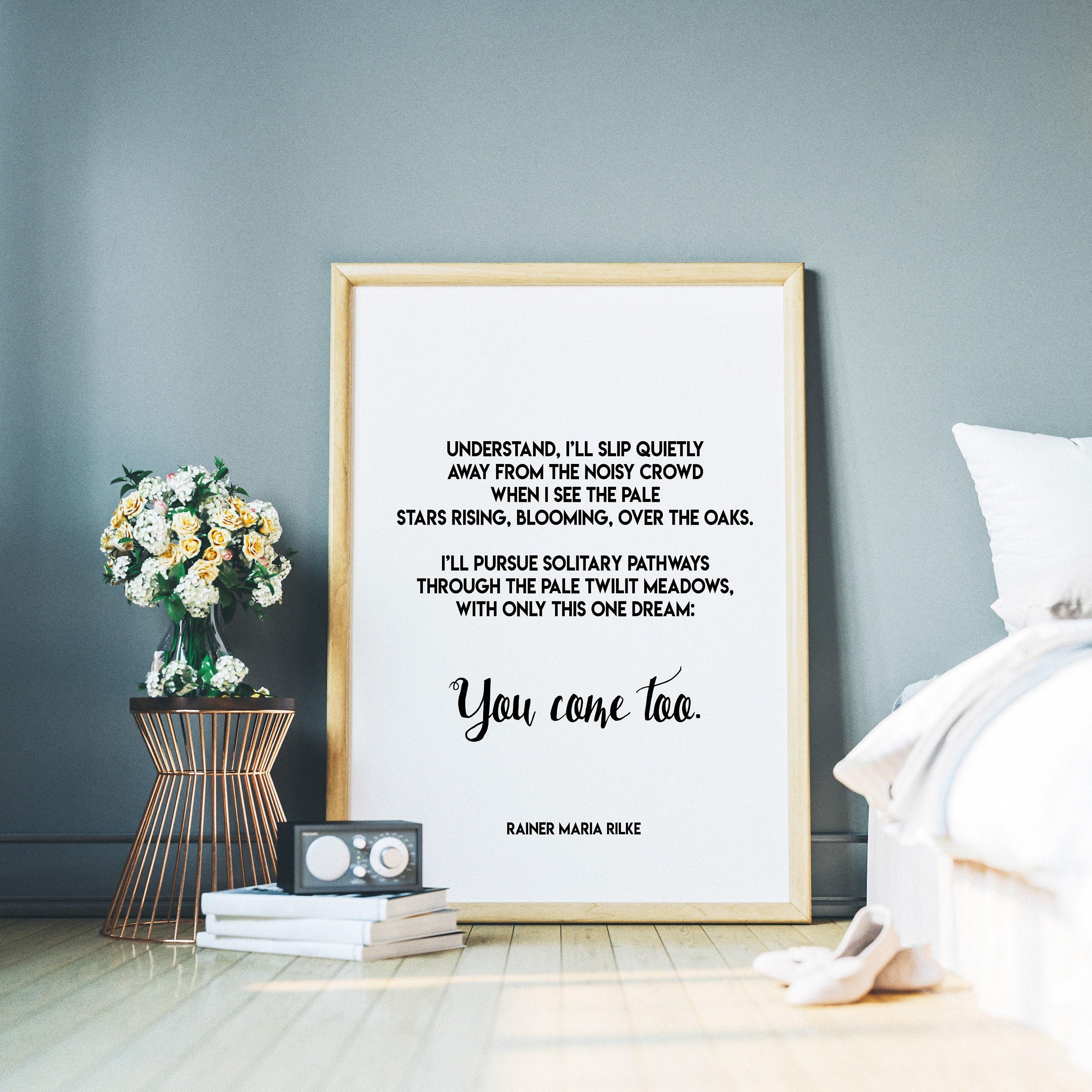 A framed print with a white background and black text, featuring a Rilke poem, leans against a gray wall, with a small table holding flowers and books to the left and a white bed to the right.
