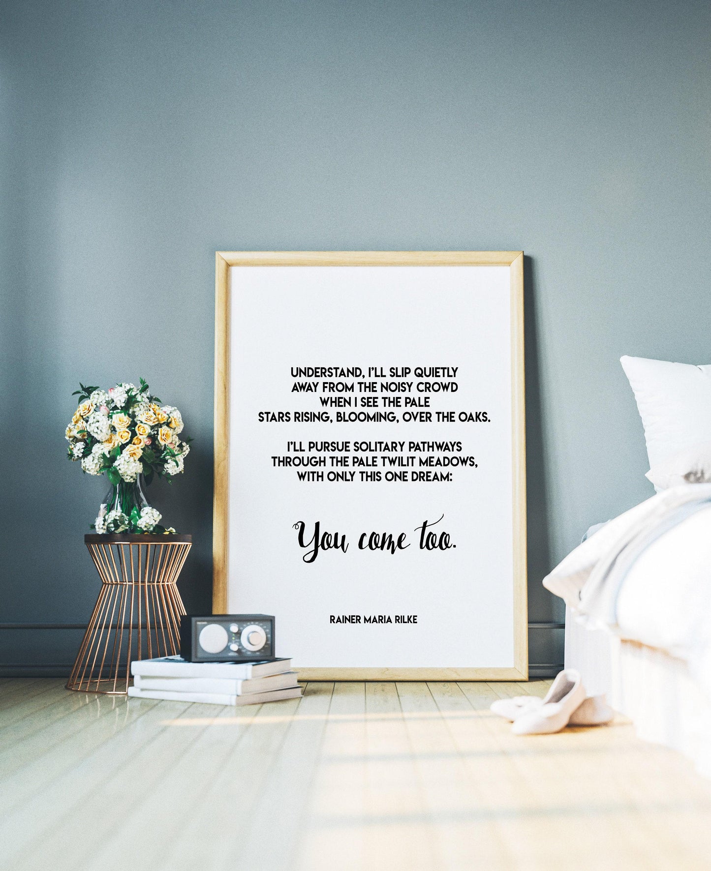 A framed print with a white background and black text, featuring a Rilke poem, leans against a gray wall, with a small table holding flowers and books to the left and a white bed to the right.