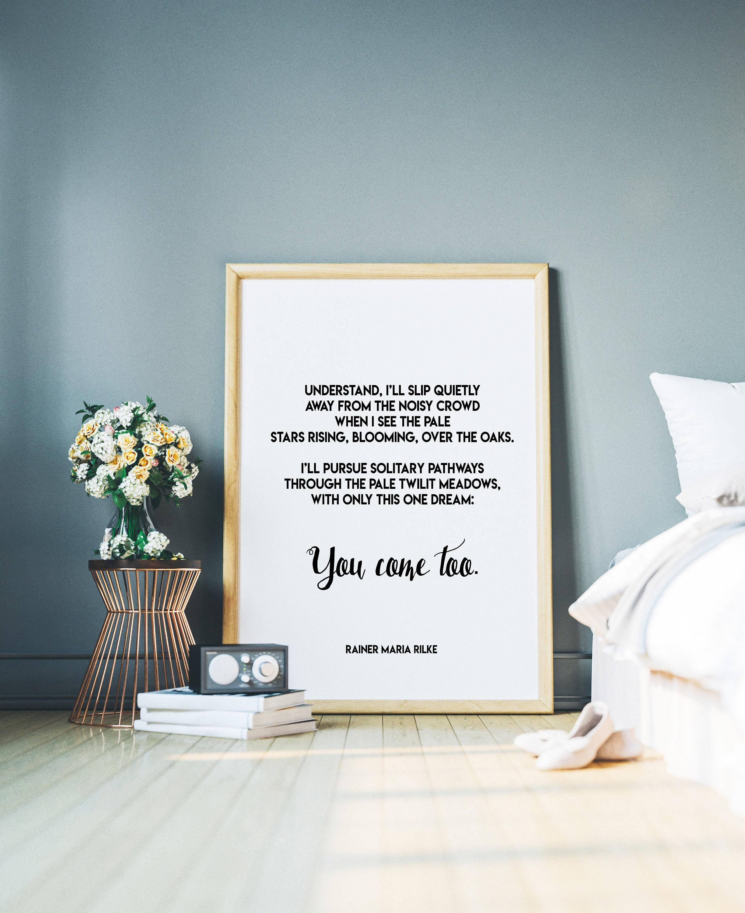 A framed print with a white background and black text, featuring a Rilke poem, leans against a gray wall, with a small table holding flowers and books to the left and a white bed to the right.