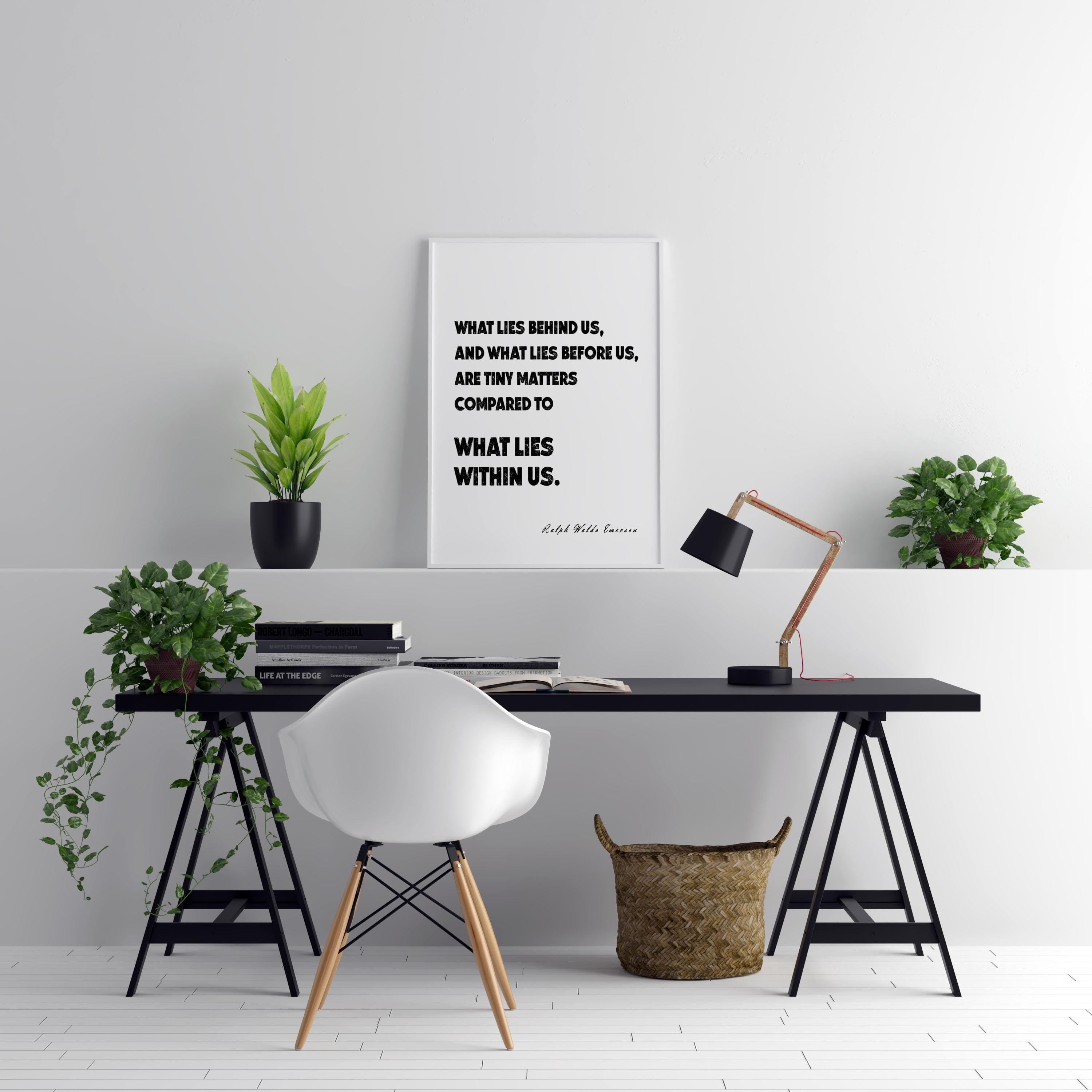 A white framed print with the quote, "WHAT LIES BEHIND US, AND WHAT LIES BEFORE US, ARE TINY MATTERS COMPARED TO WHAT LIES WITHIN US. Ralph Waldo Emerson." hangs above a black desk with a white chair and two potted plants.