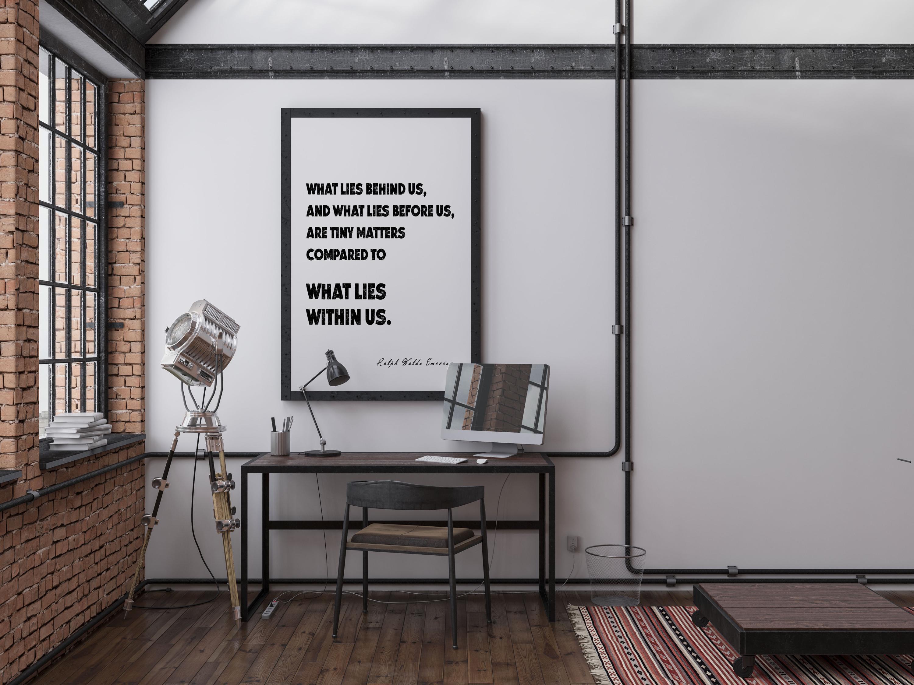 A black framed print with the quote, "WHAT LIES BEHIND US, AND WHAT LIES BEFORE US, ARE TINY MATTERS COMPARED TO WHAT LIES WITHIN US. Ralph Waldo Emerson." hangs on a white brick wall above a black desk with a computer monitor.
