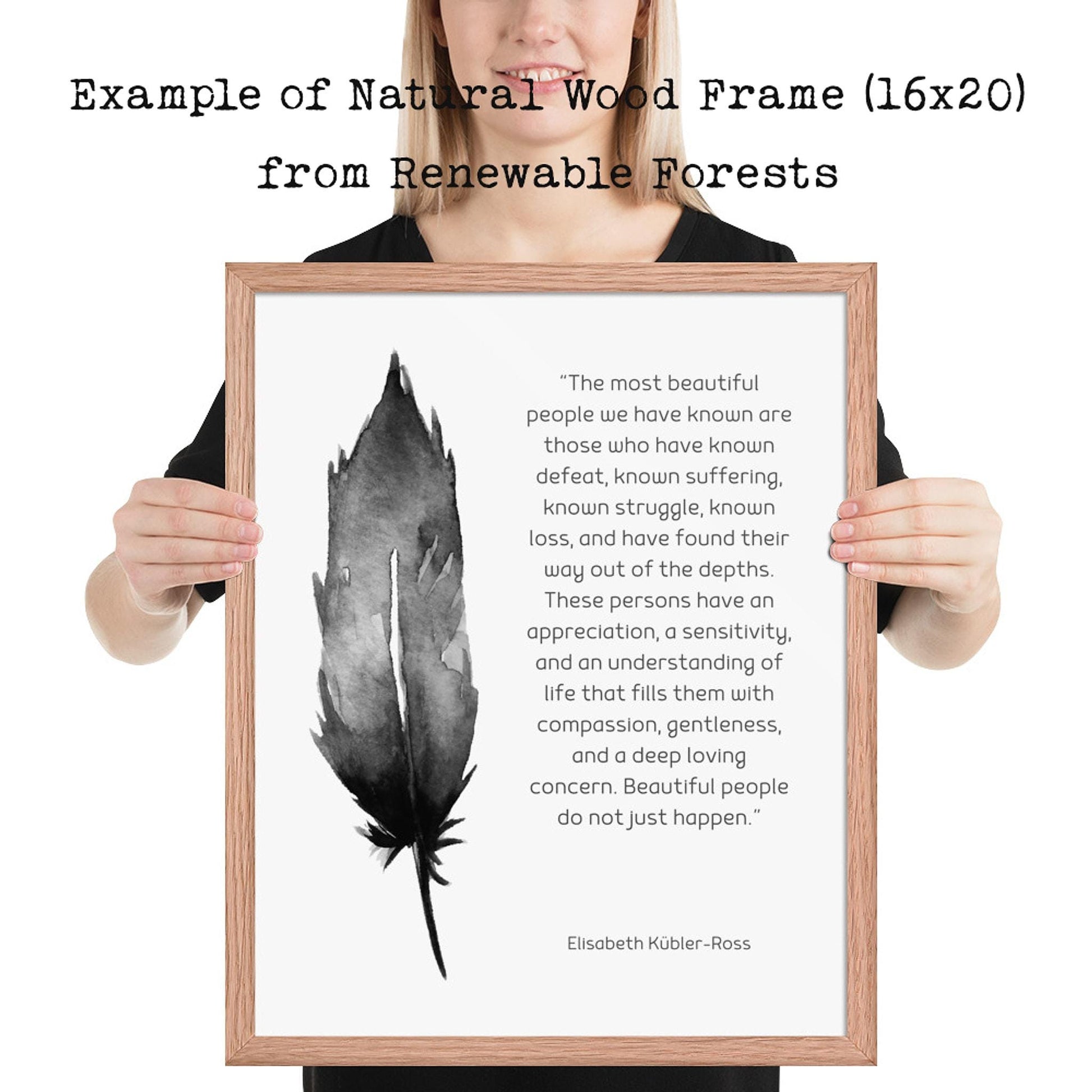 A person holds a light wood-framed print with a black feather illustration and a quote by Elisabeth Kübler-Ross on a white background.